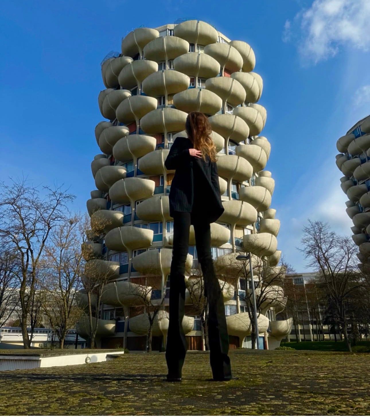 A woman in black seen from behind, standing before a residential tower with rounded, petal-shaped concrete balconies under a blue sky