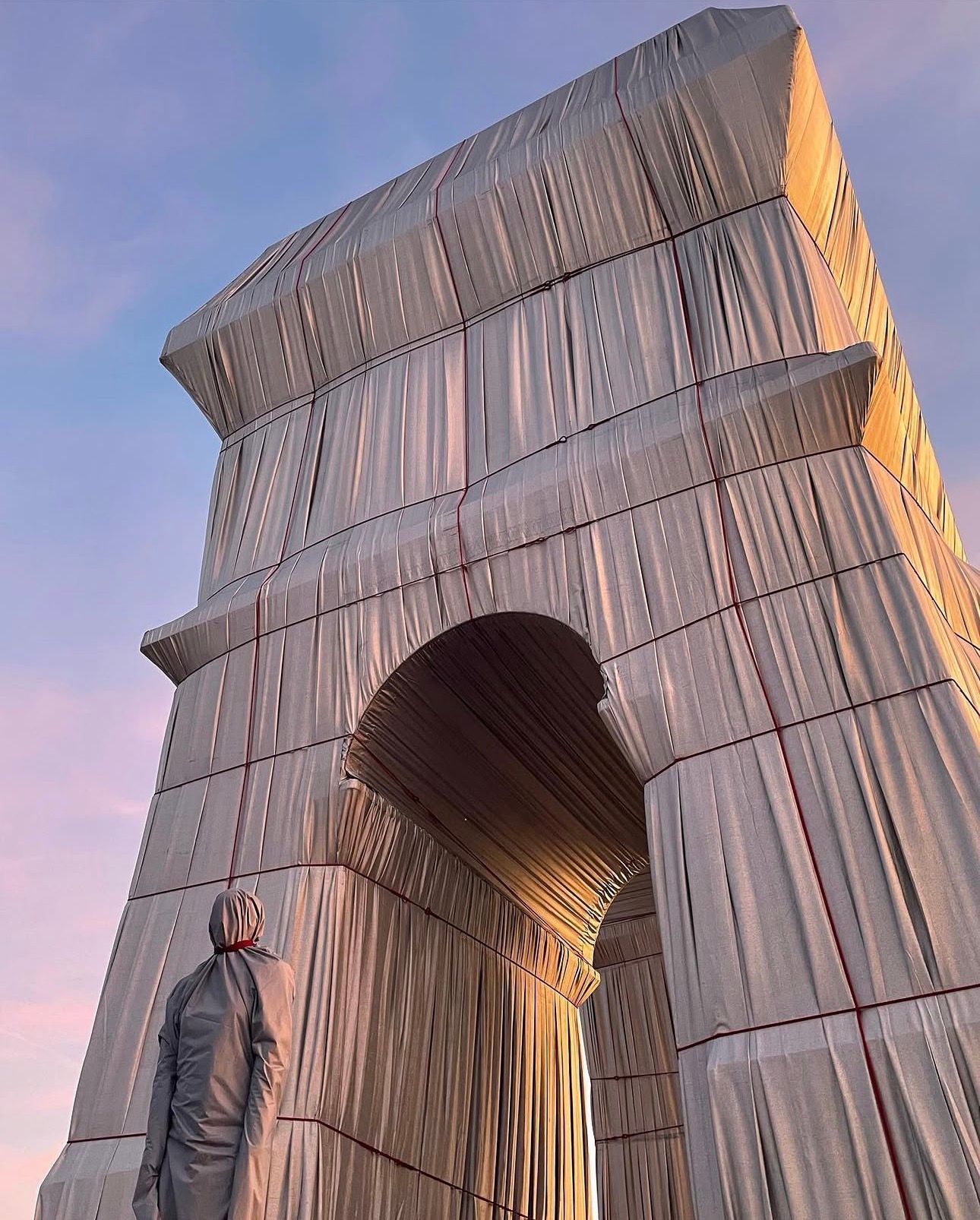 A figure in a grey hooded suit looks up at the wrapped Arc de Triomphe, its fabric folds glowing at dusk