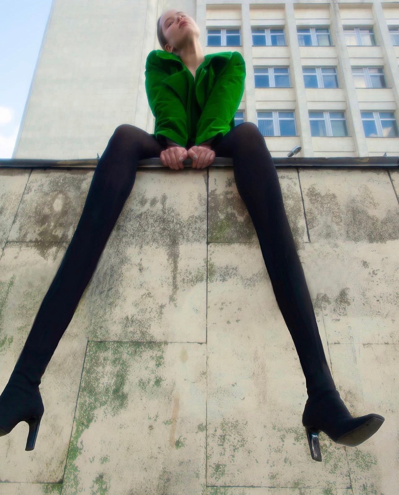 A woman in a green jacket sits on top of a weathered concrete wall, her elongated legs in black tights stretching down the wall, a modernist building behind