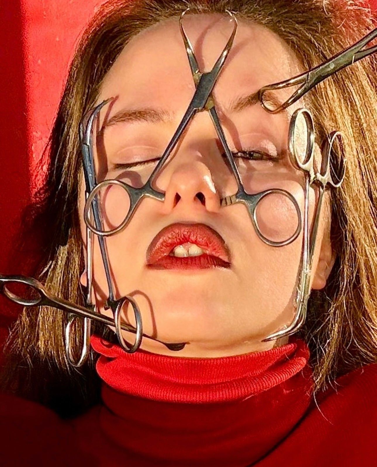 Close-up of a woman's face with eyes closed, surgical clamps resting across her face, against a red background