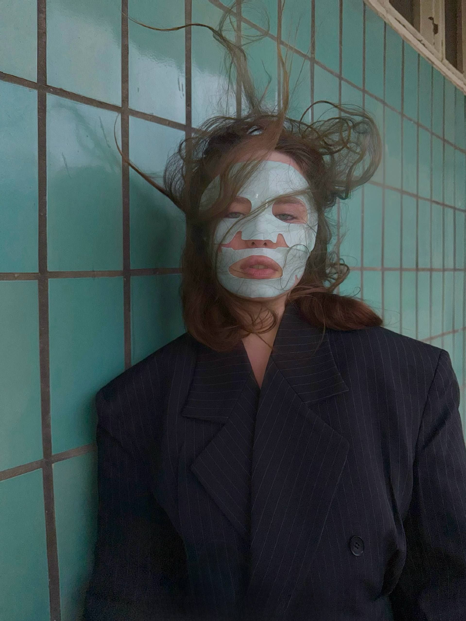 Portrait of a woman wearing a pale sheet face mask, hair blown by wind, in a pinstripe jacket against a turquoise tiled wall